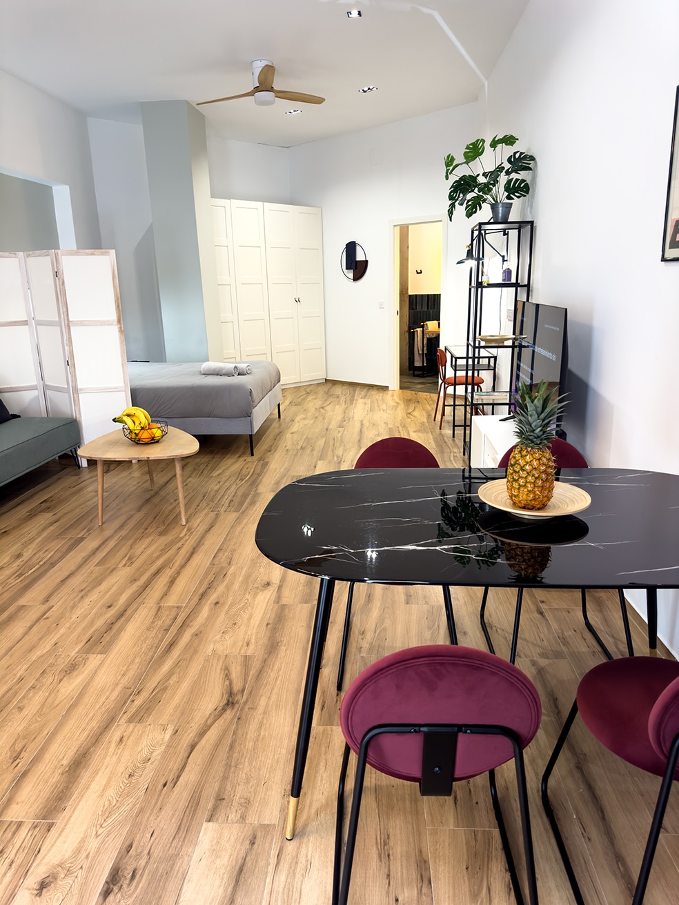 An open living space features a dark marble dining table with four burgundy chairs, set against light wooden flooring. A comfortable sofa and a queen size bed are visible in the background, complemented by green plants and a decorative mirror on the wall.
