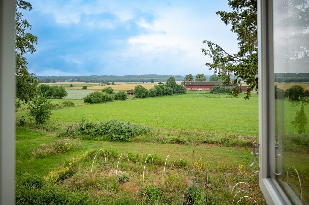 A scenic view from the window captures the expansive green fields and dotted trees, framed by the white window. Rolling landscapes extend into the distance, showcasing a classic red Swedish house nestled among the greenery, offering a serene glimpse of the natural surroundings.