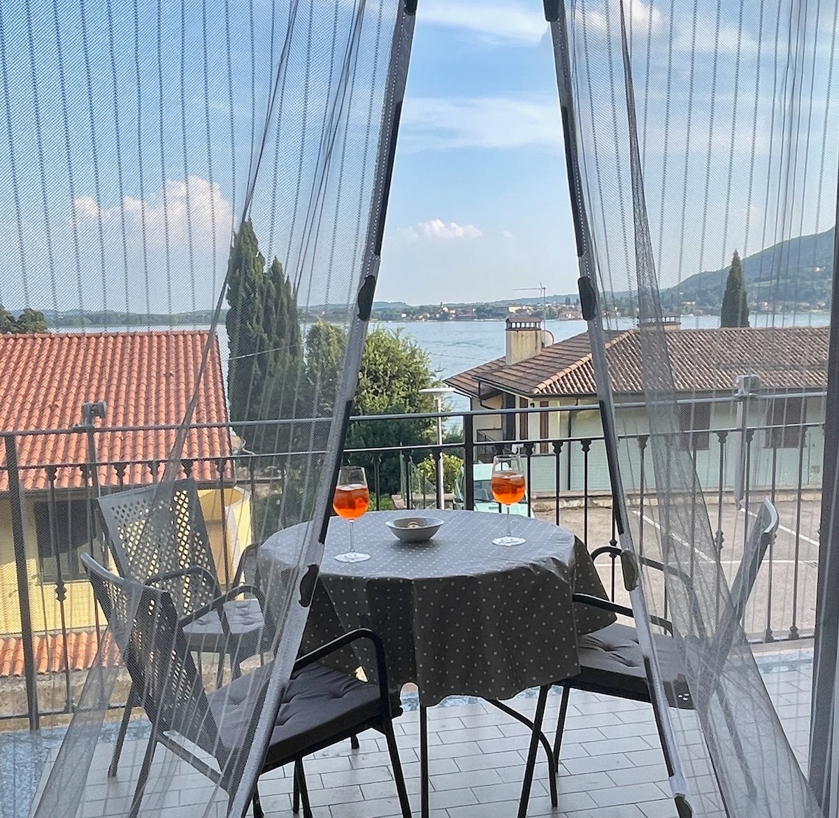 A balcony is shown with a round table covered in a dotted tablecloth, surrounded by two dark chairs. Two glasses of orange drinks are placed on the table, with a panoramic view of the lake visible through a sheer curtain.