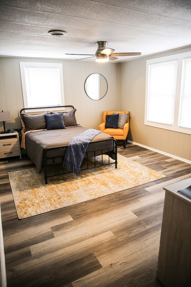 A well-lit bedroom showcases a metal bed frame with a neatly made bed, complemented by a soft throw and decorative pillows. An orange accent chair with textured cushions enhances the space. Large windows provide natural light, and a round mirror adds a stylish touch.