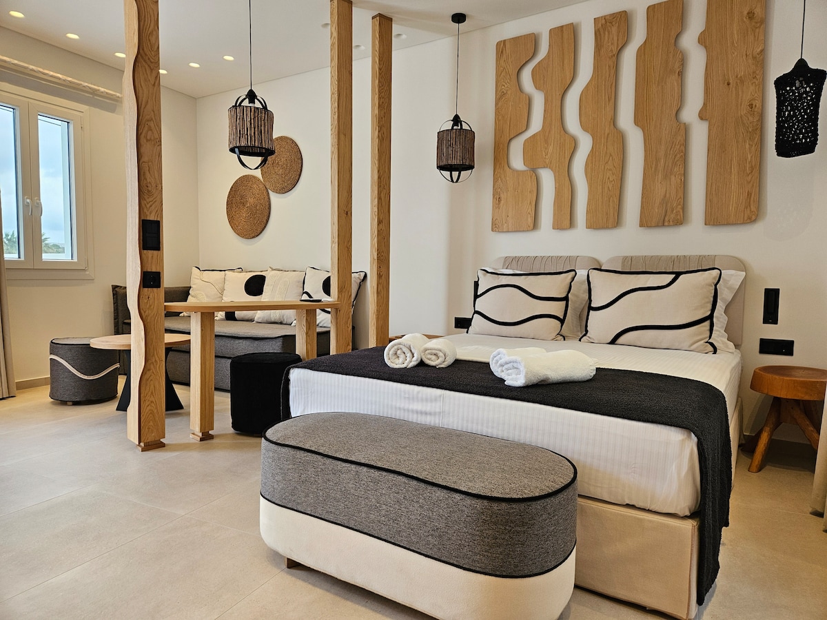 An open plan suite showcases a spacious bed with black and white bedding against wood-accented walls. A seating area is arranged with a small dining table and decorative wall art. Natural light enters through a window, creating a bright, inviting atmosphere.