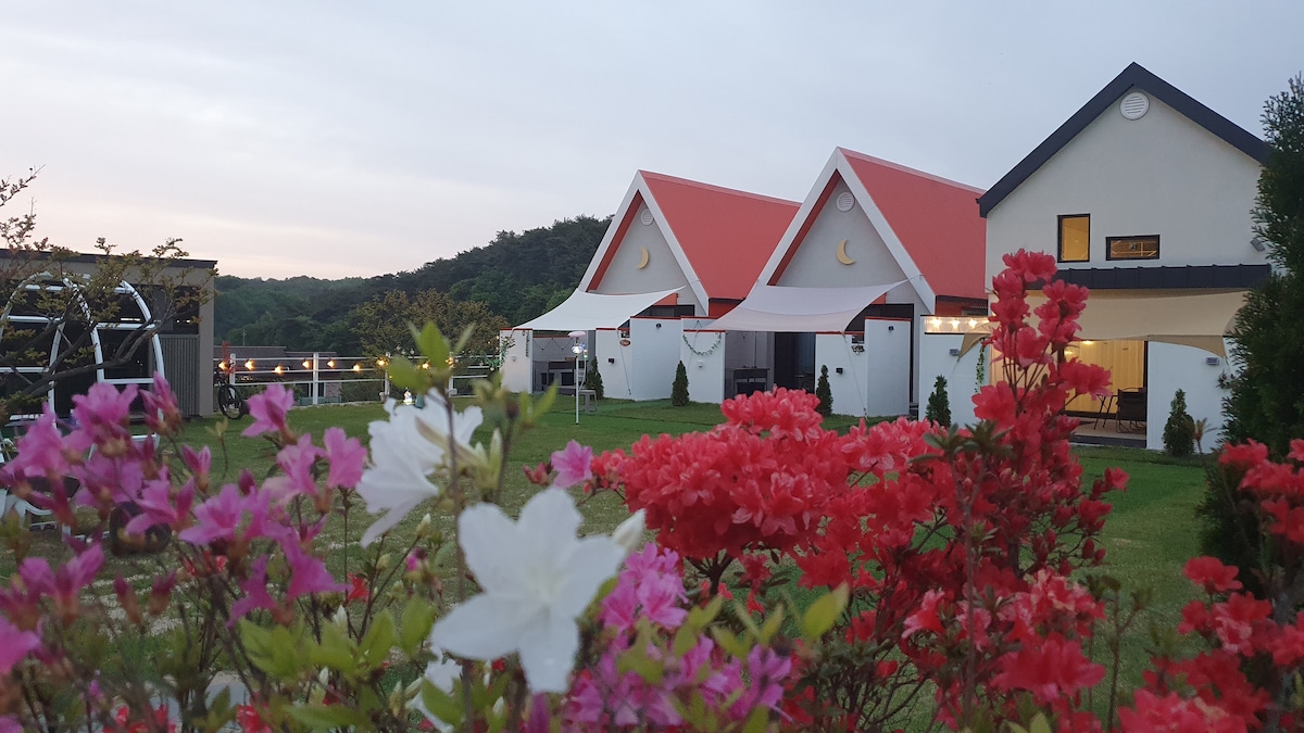 The image captures a scenic view of a tranquil house with a distinctive red roof, surrounded by vibrant flowers in white and shades of pink. The building features a clean design with large windows, set against a backdrop of lush greenery and gentle hills.
