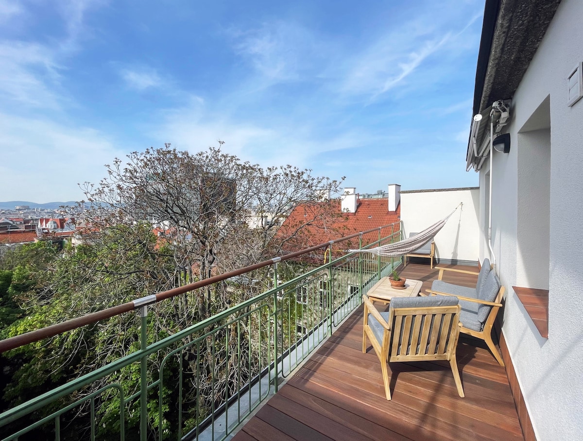 A spacious terrace features wooden decking and comfortable seating arrangements, overlooking a vibrant cityscape. A hammock is positioned for relaxation. Lush green trees are visible, adding a natural element to the urban view, while the horizon showcases rooftops and the skyline.