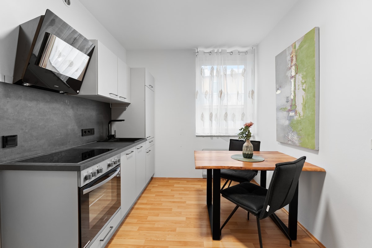 A modern kitchen area features sleek cabinets and an integrated oven. A wooden dining table is set with a vase of flowers, complemented by two black chairs. Natural light flows through the window, enhancing the grey and white color scheme.
