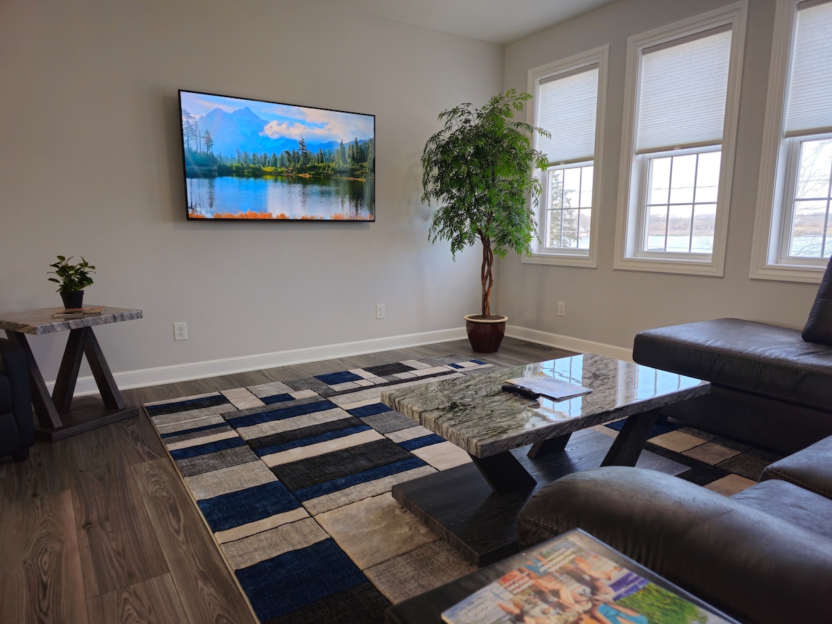 A modern living area is highlighted by a large flat-screen television displaying a serene landscape. A textured area rug with geometric patterns covers the floor, while a potted plant adds a touch of greenery. Natural light enters through two large windows.