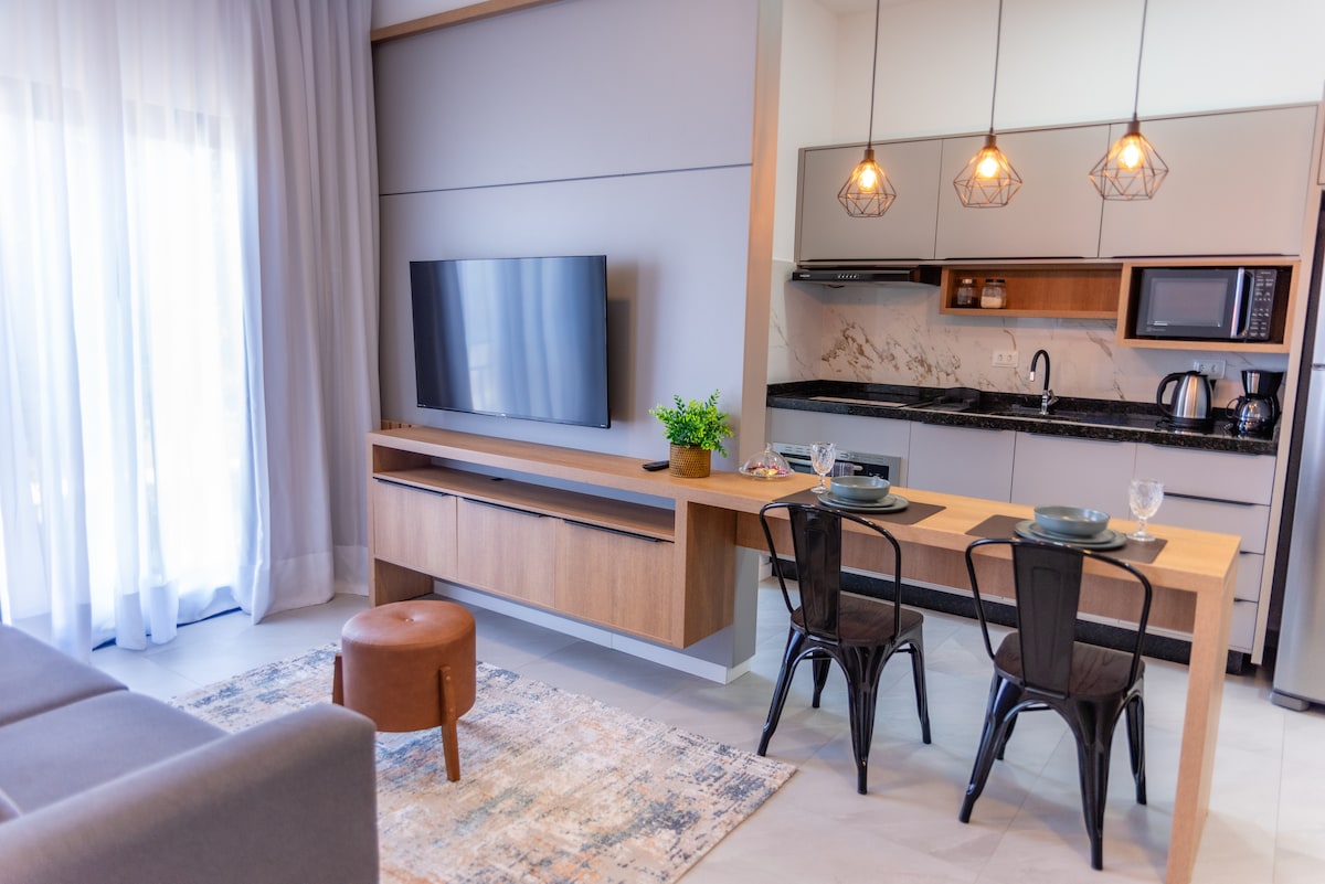 An inviting space features a modern kitchen area with sleek cabinets and a granite countertop. A dining table is set for two beside a comfortable living area, illuminated by pendant lights. Large windows allow natural light to brighten the room, complemented by sheer curtains.