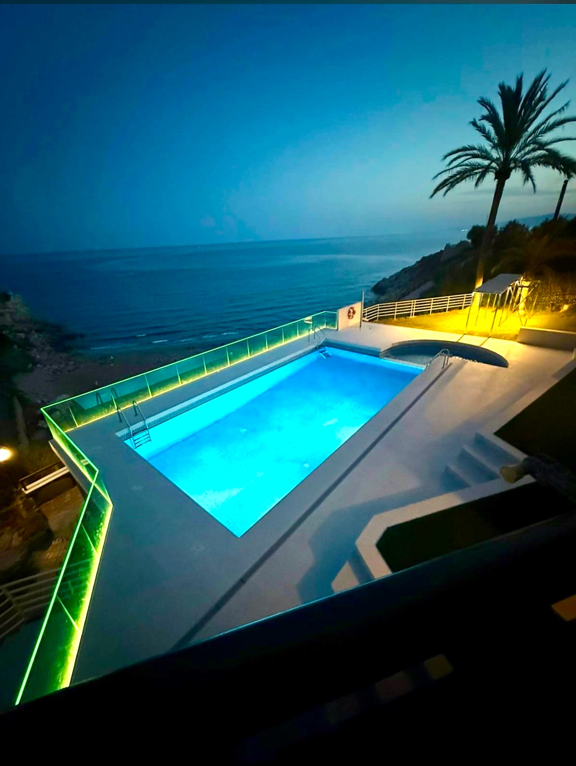 A serene pool area is illuminated in soft green lighting, reflecting off the calm water. The pool overlooks the sea, and the surrounding palm trees add a touch of tropical elegance. The scene conveys a tranquil ambiance, perfect for evening relaxation.