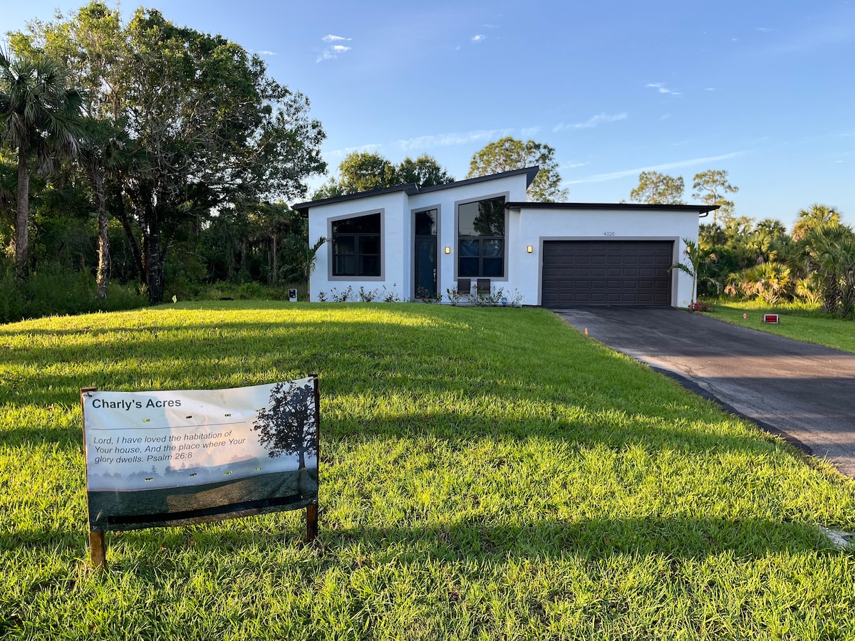 The exterior of a modern home is set against a backdrop of green grass and lush trees. A sign labeled 'Charly's Acres' is positioned in the foreground, while the home features large windows and a spacious driveway leading to a single-car garage.