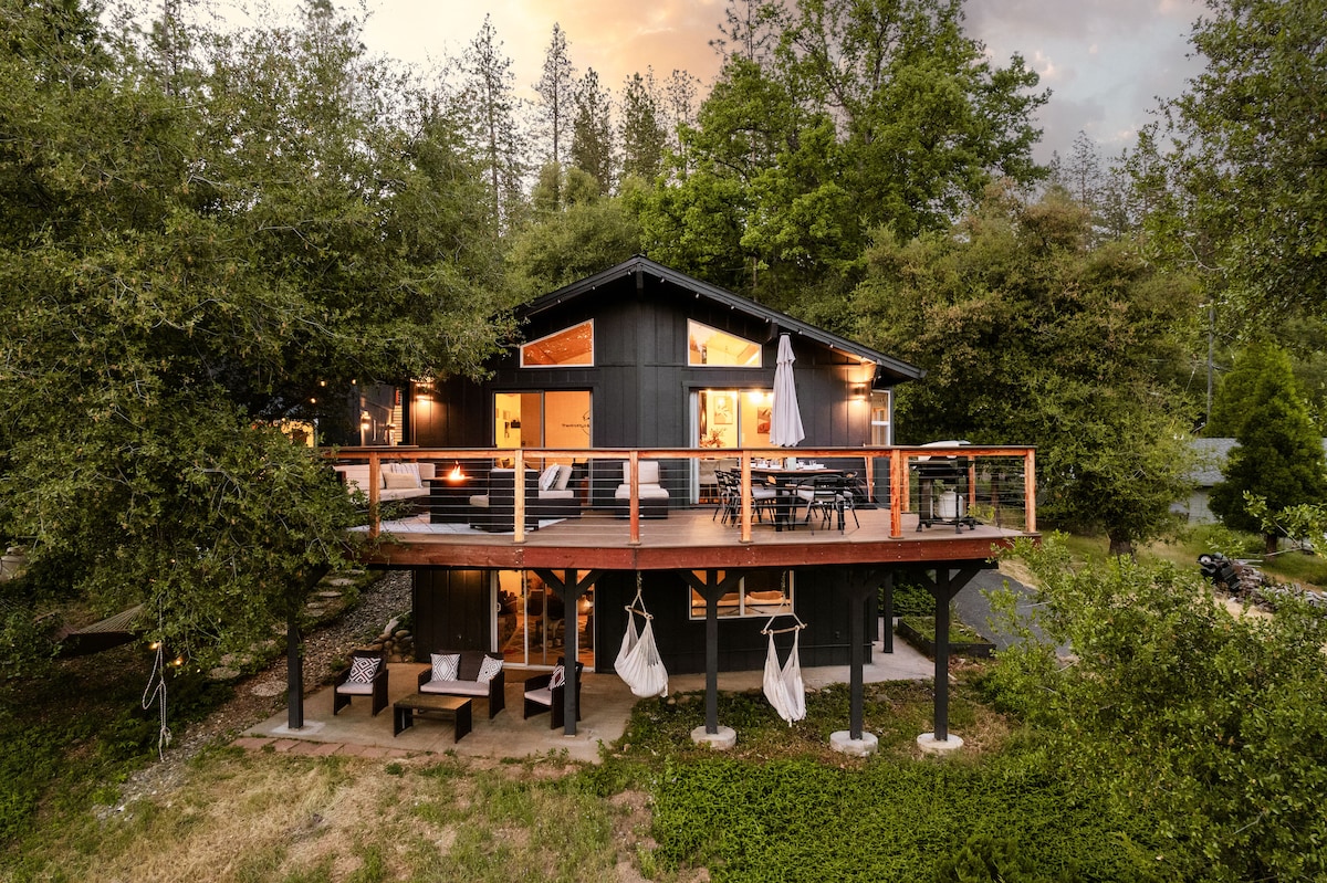 A modern black cabin is depicted, set amidst lush greenery. A spacious deck is visible, featuring outdoor seating and an umbrella. The home is accentuated by warm lighting from the windows, creating a welcoming atmosphere as dusk approaches.