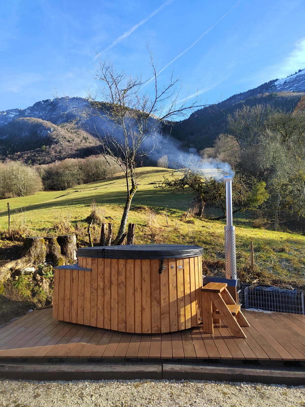 An outdoor wooden hot tub is situated on a wooden deck, surrounded by scenic mountain views. Steam rises softly from the tub, contrasting against the clear blue sky and lush green landscape, which features rolling hills and distant trees.