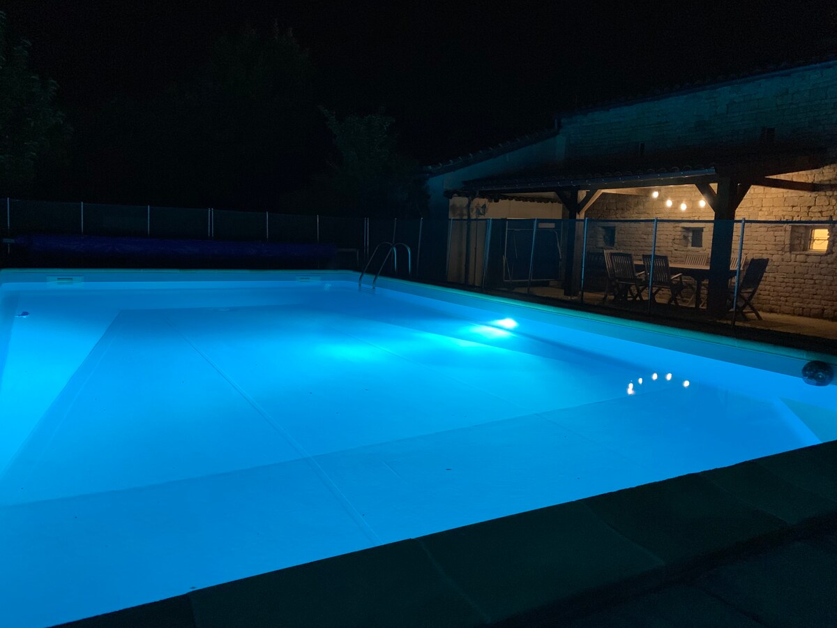 The illuminated pool reflects a serene blue at night, creating a calming atmosphere. Soft lighting from nearby outdoor seating highlights the covered area, inviting relaxation. The surrounding fence adds privacy, ensuring an exclusive and secure environment for enjoyment.