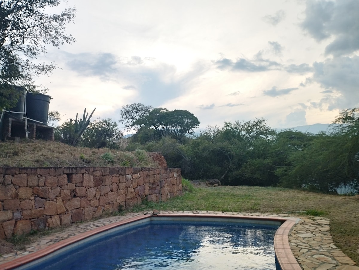 A tranquil pool is framed by a stone patio and bordered by lush greenery. The surrounding landscape features low shrubs and trees under an expansive sky with gentle cloud formations, creating a peaceful outdoor setting.