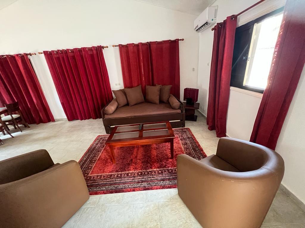 A comfortable living area features a brown couch with matching cushions, accompanied by two round armchairs. A wooden coffee table sits on a decorative red rug. Rich red drapes frame the window, allowing natural light to gently illuminate the space.