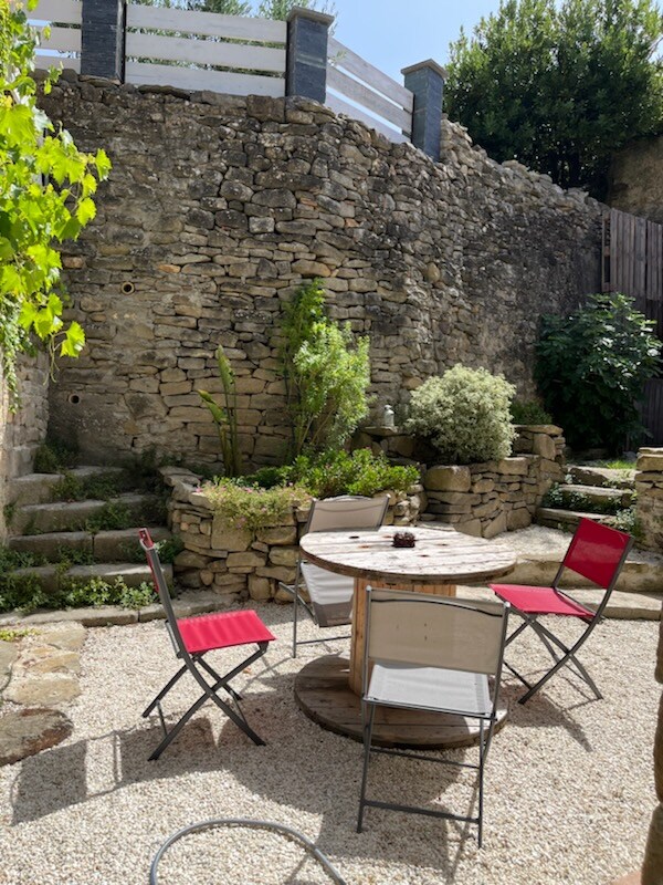 An intimate garden area features a circular wooden table surrounded by four chairs in contrasting colors. Stone walls and steps create a rustic backdrop, while greenery adds a touch of natural beauty. The space is designed for relaxation and quiet enjoyment.