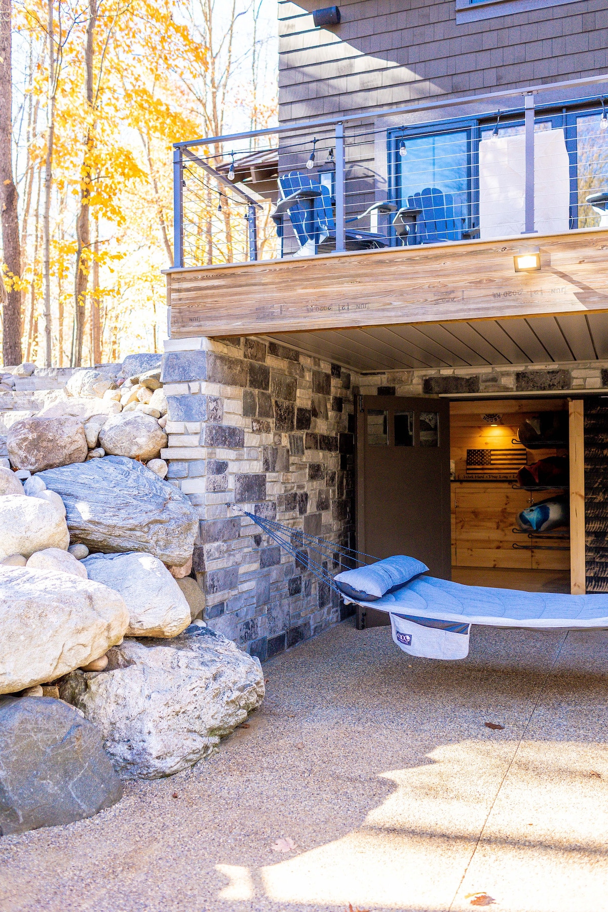 A stone wall with natural boulders borders the outdoor area, leading to a covered space fitted with a hammock and storage. Above, a deck is visible with outdoor furniture, framed by trees showcasing autumn foliage. The entrance features a simple door against wooden paneling.