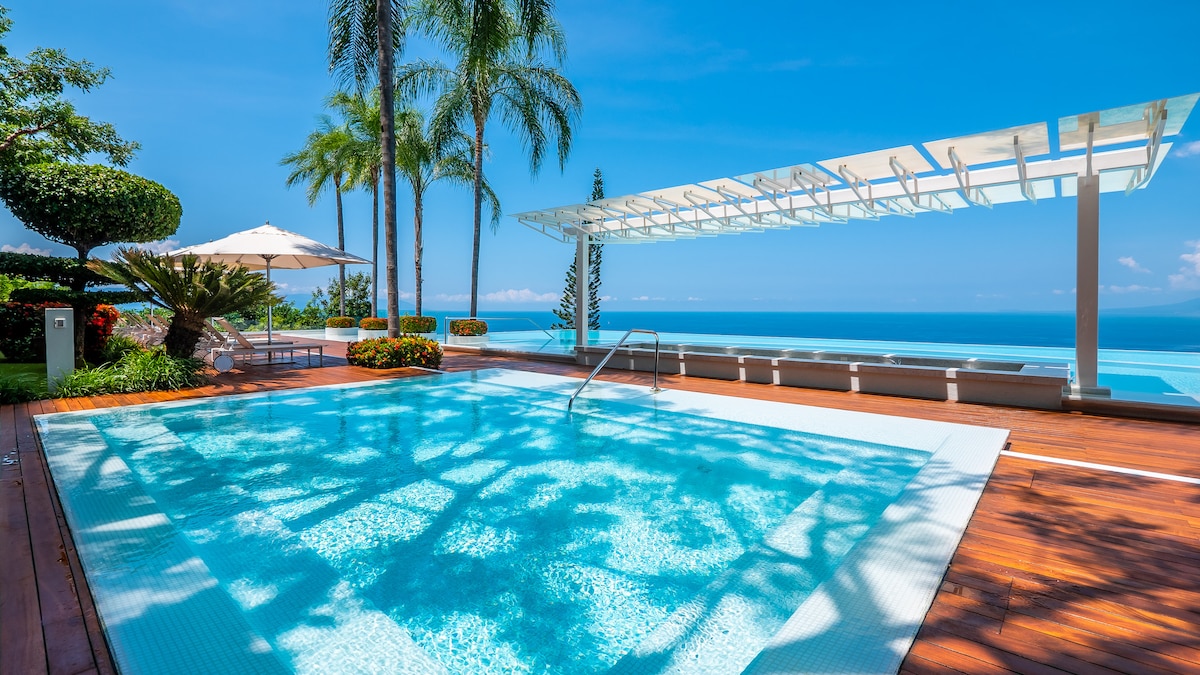An inviting private pool is surrounded by lush greenery and palm trees, reflecting the clear blue sky. Comfortable sun loungers are positioned nearby, providing a perfect spot for relaxation, while a shaded area with umbrellas enhances the outdoor space.