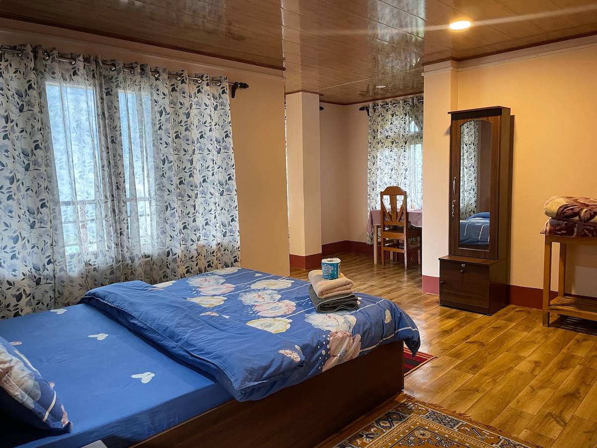 A spacious room features a large bed dressed with a floral blue coverlet. Natural light filters through two windows adorned with patterned curtains. A wooden dresser stands against one wall, accompanied by a dining table and chair set in the background.