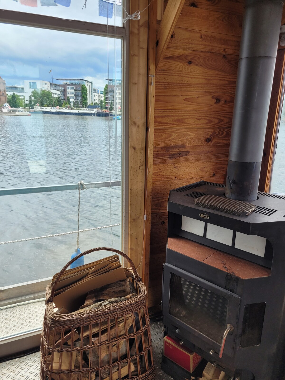 A cozy corner features a wood stove with a metal chimney, positioned next to a large window offering views of the water. A woven basket filled with logs is placed nearby, against wooden paneling that enhances the rustic feel of the space.