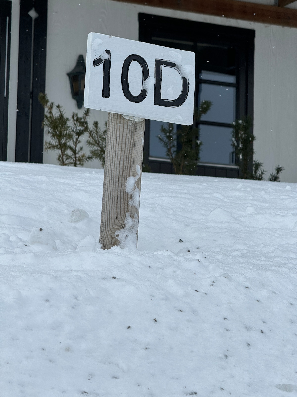 A white wooden sign displaying the number '10D' stands upright in a snow-covered landscape. The sign is surrounded by a blanket of fresh snow, with only a hint of greenery visible in the background and light flakes of snow gently falling.