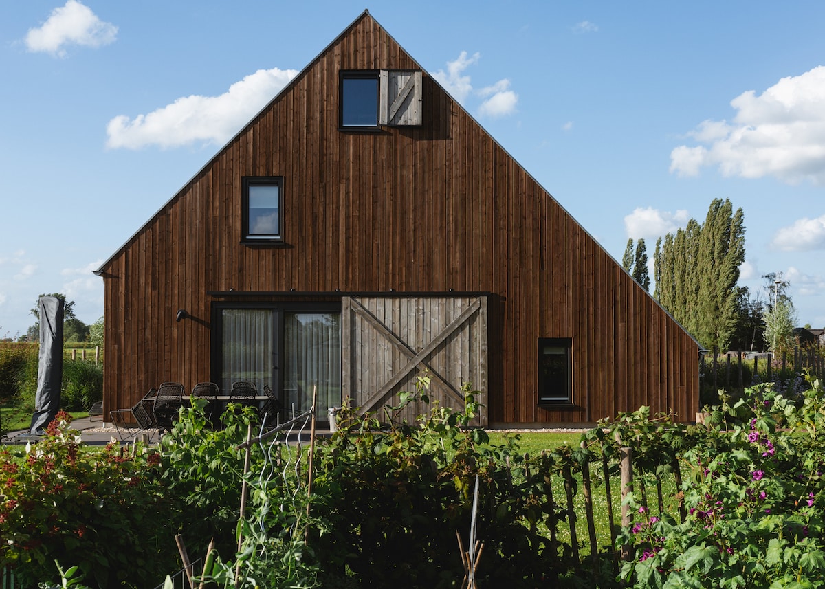 The exterior of the lodge features a striking wooden façade with a steep gabled roof. Large glass doors provide access to the outdoor area, while a charming garden filled with greenery and flowers sits in the foreground, enhancing the lodge's natural setting.