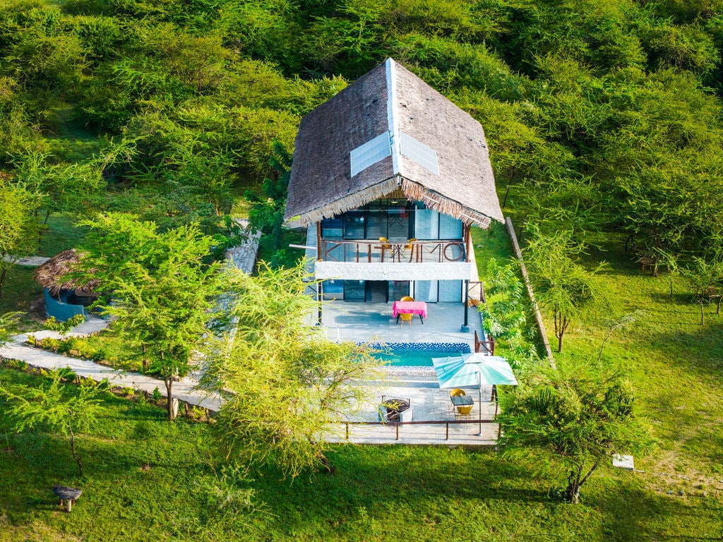 An aerial view of a two-story lodge nestled among lush greenery. The structure features a thatched roof and expansive windows, with a private pool located on a spacious patio. Surrounding paths lead through the vibrant landscape, creating a serene outdoor setting.