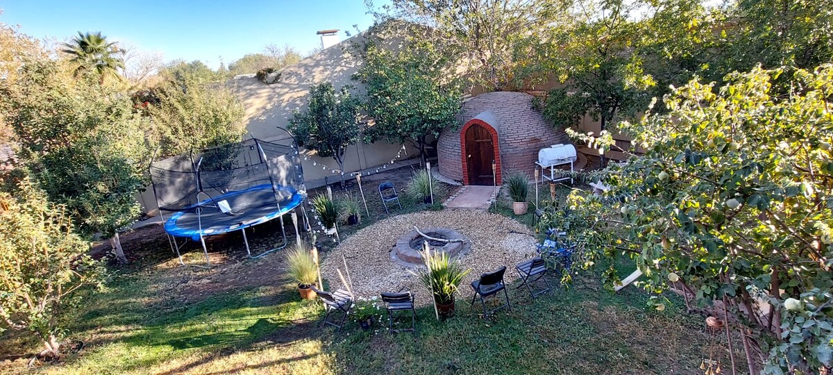 A spacious outdoor area features a circular stone fire pit surrounded by several chairs. A trampoline is visible in one corner, while lush greenery frames the space. A rustic structure with an arched door adds character, and shaded seating areas are present throughout the yard.