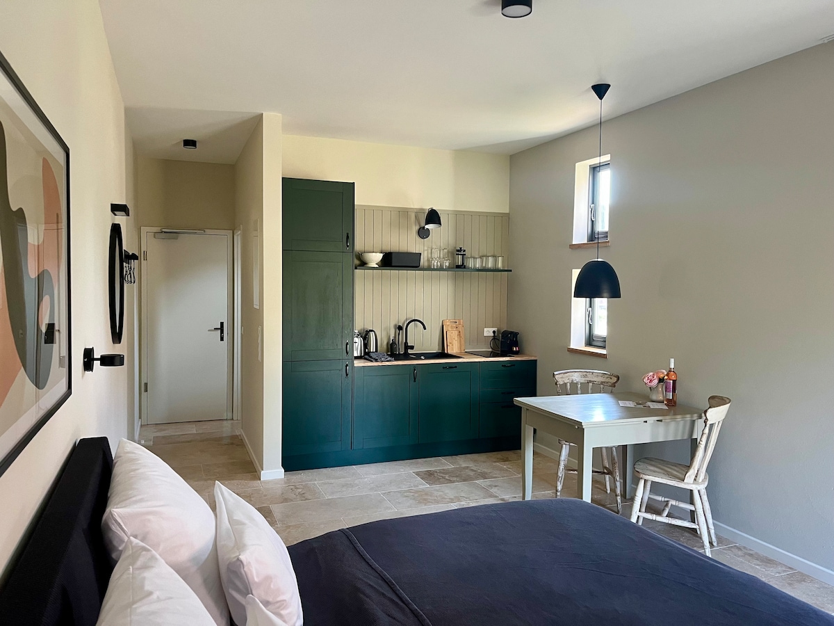 An open living and sleeping area is captured, featuring a spacious bed with crisp linens. A kitchenette with dark green cabinetry and essential appliances is visible, alongside a dining table and a couple of chairs, enhancing the room's functionality and inviting atmosphere.