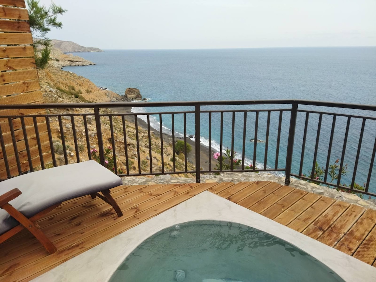 A wooden deck features a small jacuzzi, surrounded by a railing. In the background, unobstructed views of the sea and coastline are visible. A single lounge chair is positioned beside the jacuzzi, providing additional space for relaxation.
