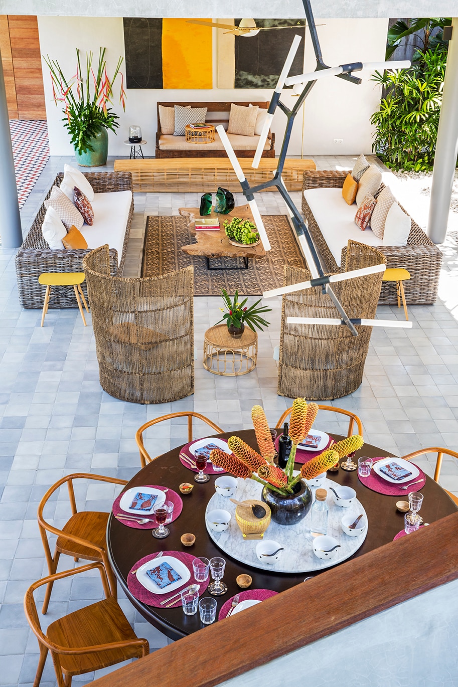 A semi-open living space is featured, showcasing a circular dining table set with colorful plates and glasses. Comfortable sofas with cushions are positioned around the central area, complemented by decorative plants and artistic lighting fixtures. Lush greenery is visible in the background.