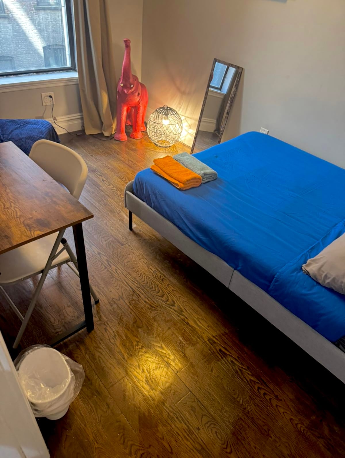 A bright private room features a spacious bed covered with a blue blanket and neatly arranged towels. A small wooden table with chairs is visible, alongside a decorative elephant figure and a light source. Natural light enters through the window, illuminating the wooden floor.