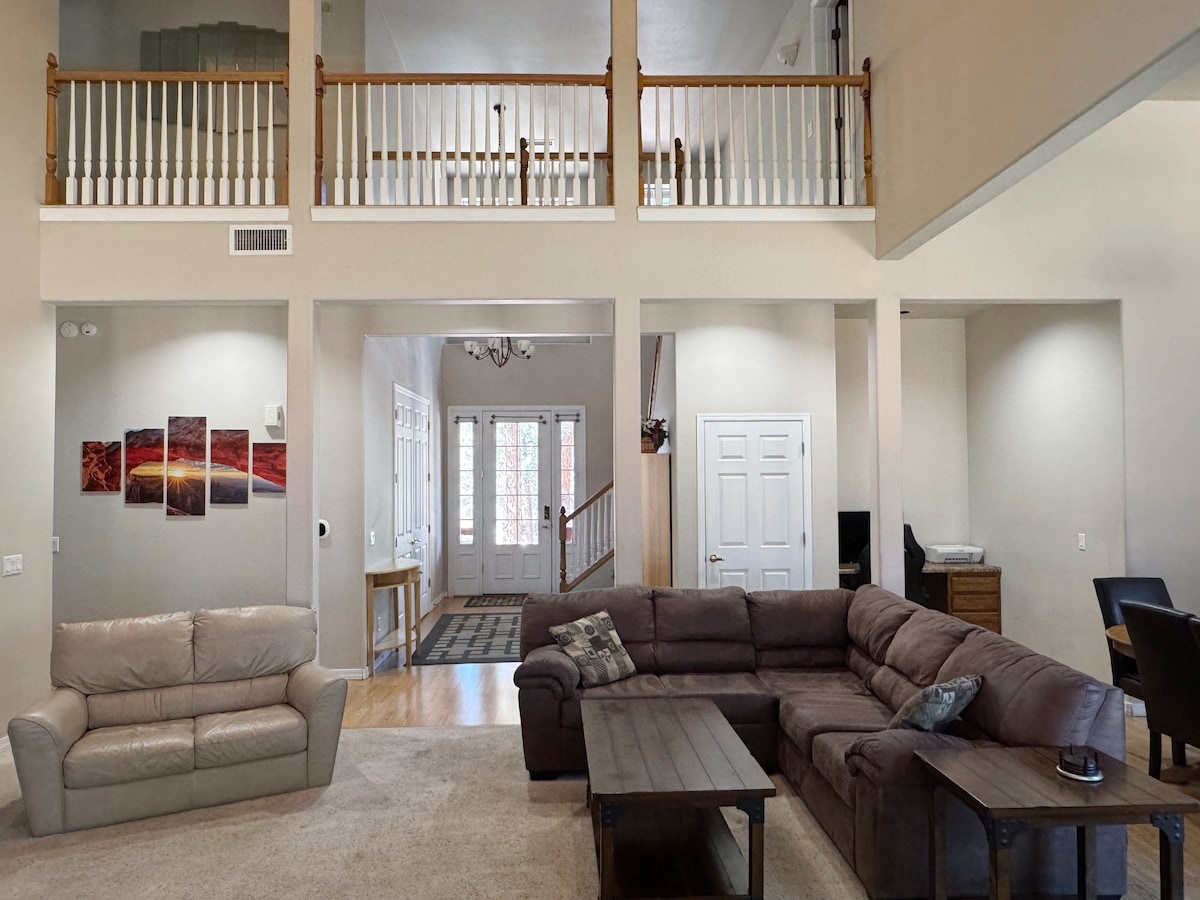 An open living area showcases a large sectional sofa arranged around a dark wooden coffee table. Light wood flooring extends through the space, leading to a staircase in the background. Large windows allow natural light to brighten the room, contributing to a welcoming atmosphere.