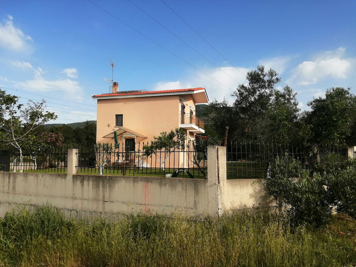 A two-story villa is set within a spacious garden, surrounded by grass and trees. The house features a light-colored facade with balconies on the upper level. A secure fence encloses the property, offering privacy and tranquility in a serene, natural environment.