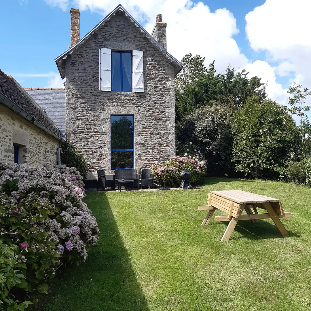 The exterior of a charming stone house is surrounded by well-maintained greenery and flowering shrubs. A wooden picnic table is placed on the lawn, providing a space for outdoor gatherings. Bright blue skies contrast with the rustic facade, highlighting the tranquil ambiance of the property.