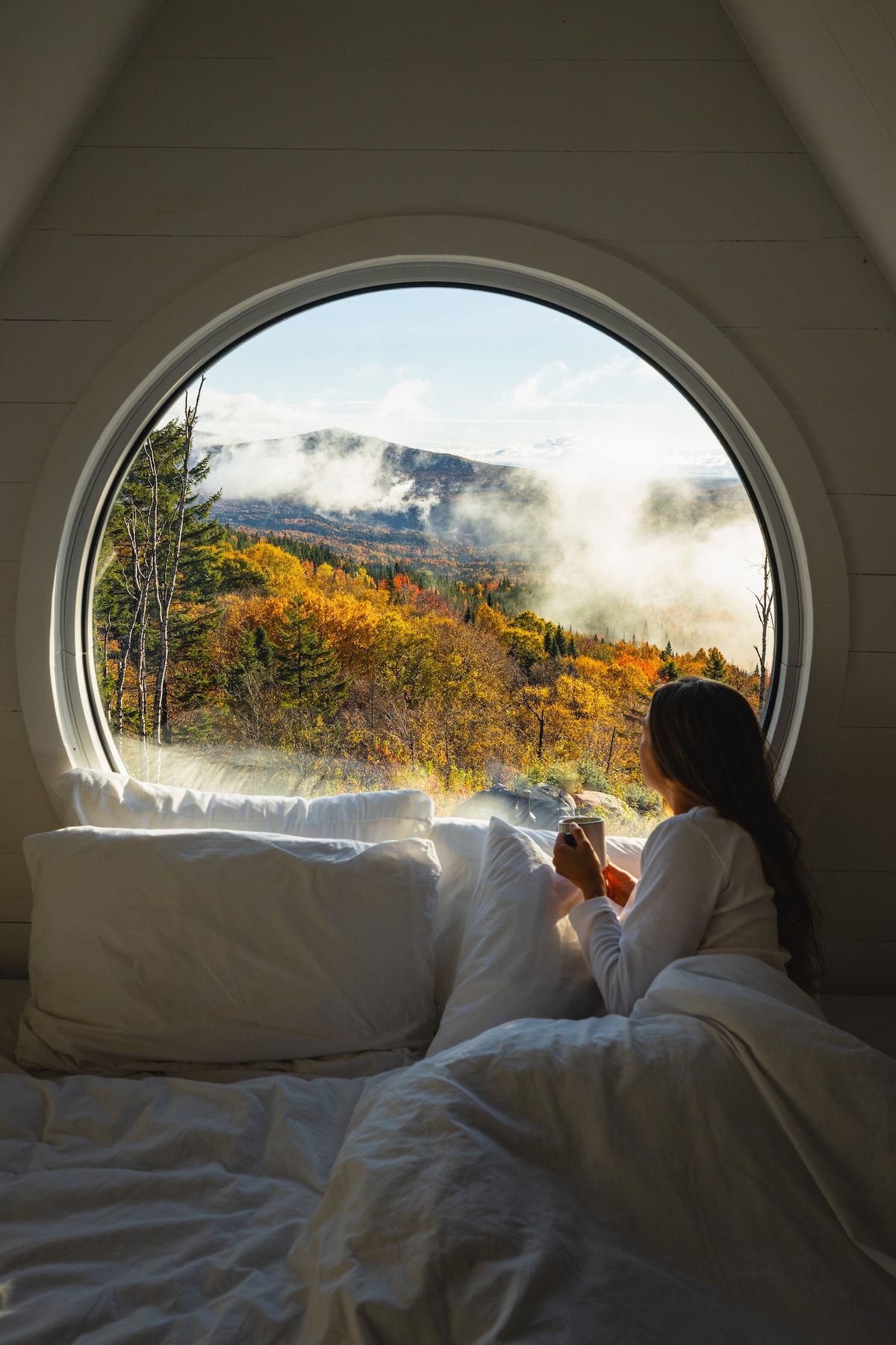 A round window frames a stunning view of vibrant autumn foliage and misty mountains. A person is seen relaxing on a spacious bed, holding a warm drink, with layers of soft white bedding providing comfort against the serene backdrop.