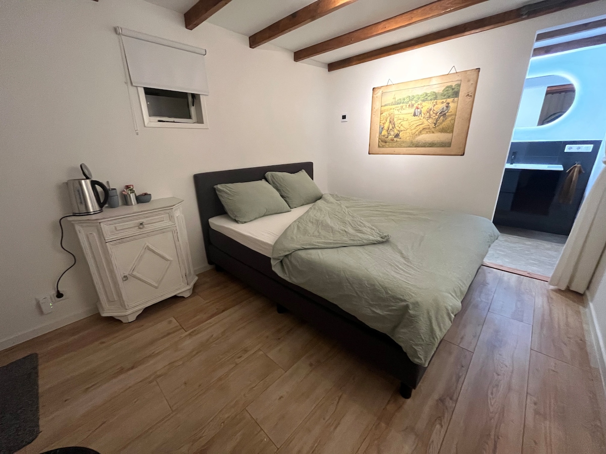 A spacious sleeping area features a neatly made bed with light green bedding and two soft pillows. A nightstand accompanies the bed, with a kettle positioned on top. The room is illuminated by natural light from a nearby window, and wooden beams add a touch of character.