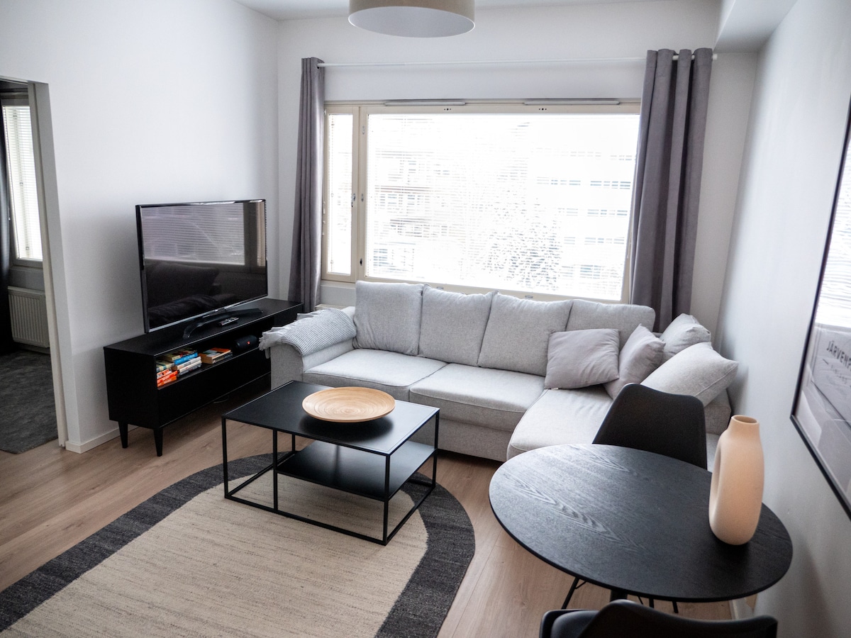 A light-filled living room features a spacious gray sofa arranged around a black coffee table. A flat-screen television is mounted on the wall, and large windows allow ample natural light. Nearby, a round dining table is set with stylish black chairs, enhancing the modern feel.