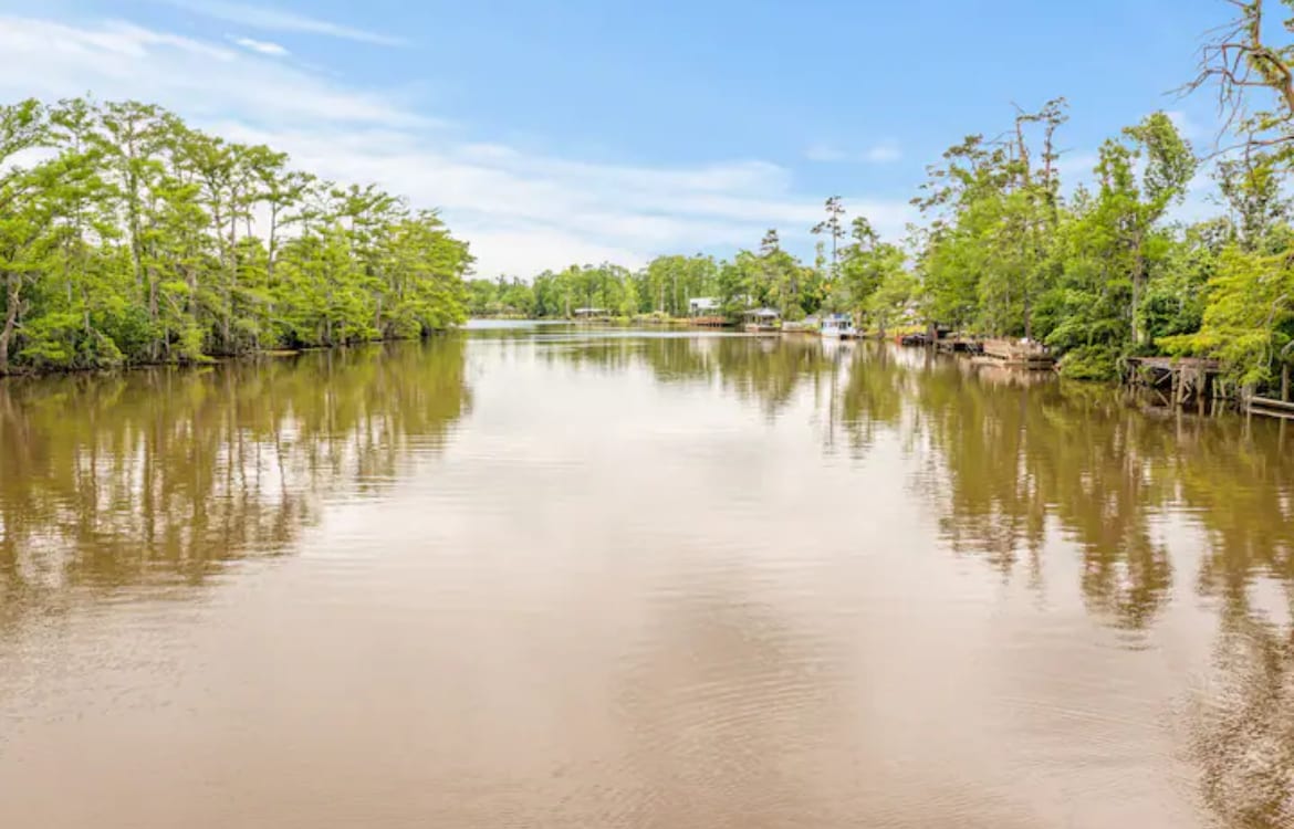 A calm river reflects the clear blue sky, bordered by lush greenery. Tall trees and occasional boats are visible along the banks, creating a serene landscape perfect for relaxation and enjoying nature.
