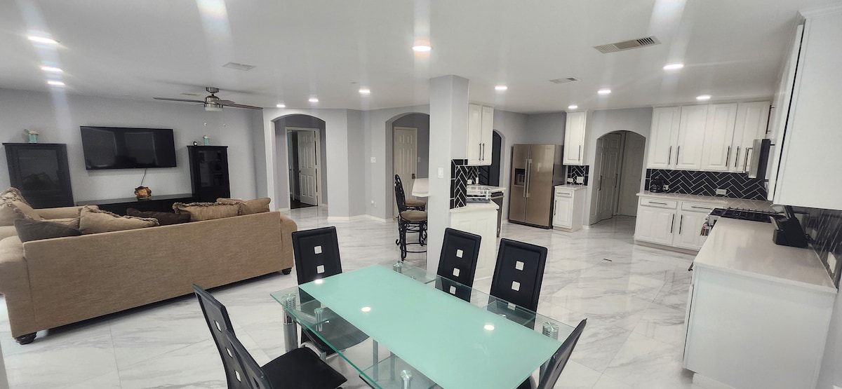 An open-concept living area features a spacious seating arrangement with a large sectional sofa facing a wall-mounted television. A modern dining table with sleek chairs is positioned nearby, with a well-equipped kitchen visible in the background, showcasing stainless steel appliances and dark cabinetry.