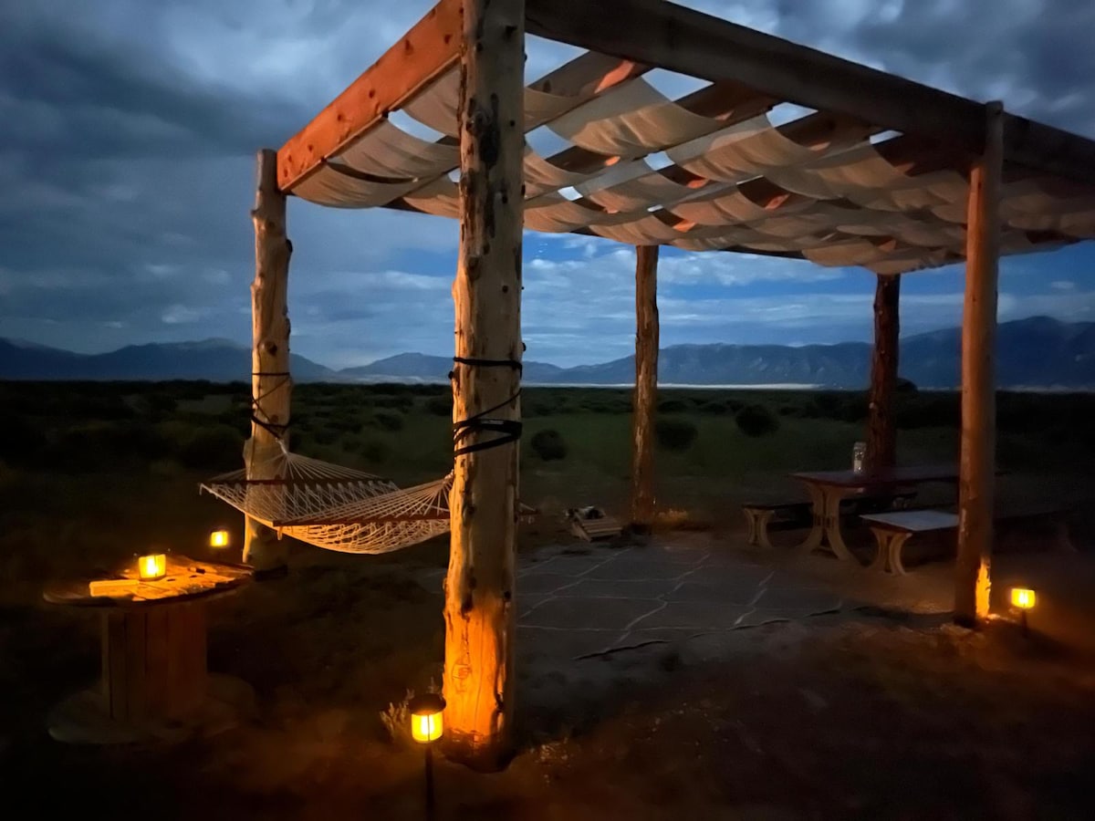 A shaded outdoor seating area is illuminated by soft lantern light, featuring a hammock suspended between wooden posts. The landscape displays distant mountain views under an evening sky, with gentle clouds adding a serene ambiance to the scene.