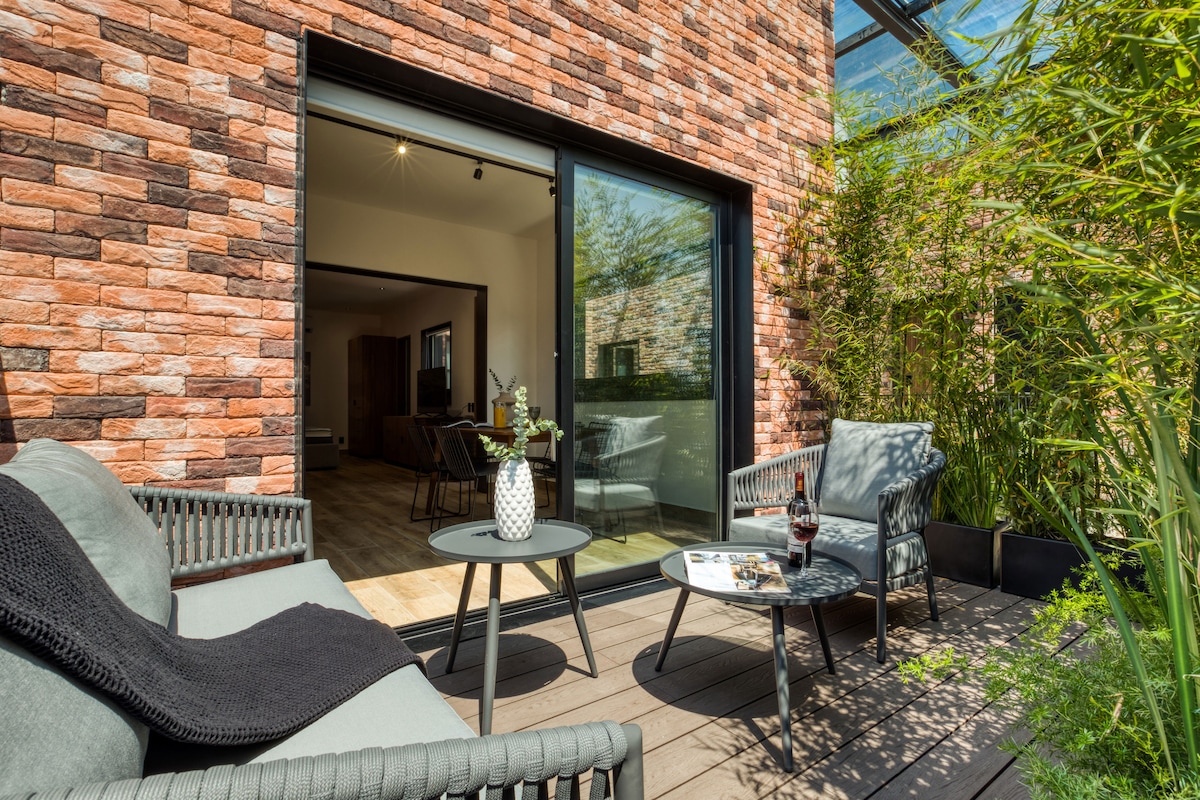 A private terrace is presented, featuring two lounge chairs and a small table surrounded by greenery. Large glass doors provide access to the interior, allowing natural light to flow into the space. The brick exterior enhances the contemporary design of the area.