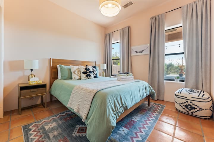 Bright bedroom with a desert-chic vibe! Plush bed with soft bedding, terracotta floors, and a patterned rug. Large windows offer backyard scenic views, while a cozy pouf and warm lighting create a relaxing space. Ideal for a serene getaway!