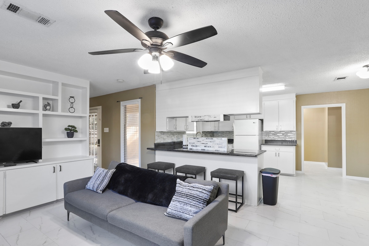 An open concept living area showcases a modern kitchen with white cabinetry and a black countertop. A comfortable sofa with decorative pillows faces a white entertainment unit holding a television. Two bar stools are positioned nearby, and light streams in through a window, enhancing the bright ambiance.