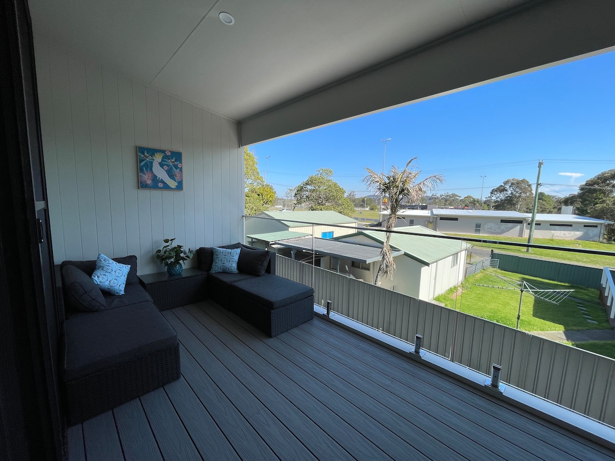 A modern balcony area featuring a comfortable seating arrangement with plush cushions. The deck is lined with smooth wooden boards, overlooking a green grassy area and surrounding rooftops. Clear skies and sunlight create an inviting atmosphere for relaxation.