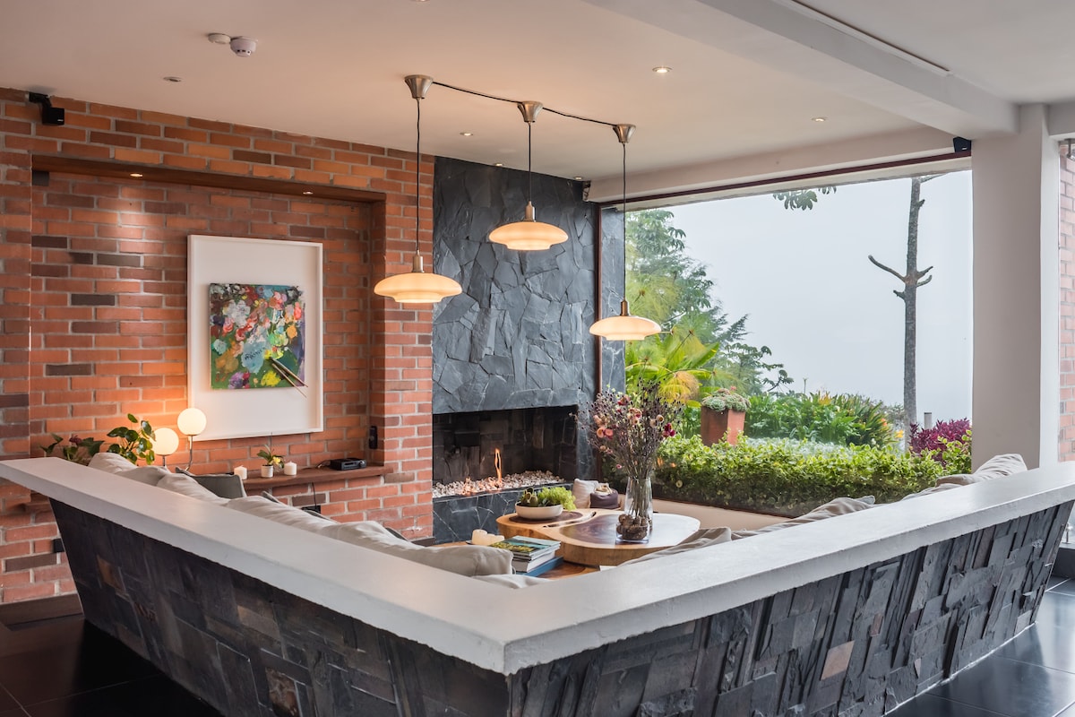 A modern living area features an angular seating arrangement with neutral-toned cushions, positioned around a round coffee table. Natural light streams through large windows, illuminating the space. A cozy fireplace is integrated into the stone wall, complemented by decorative art and greenery.
