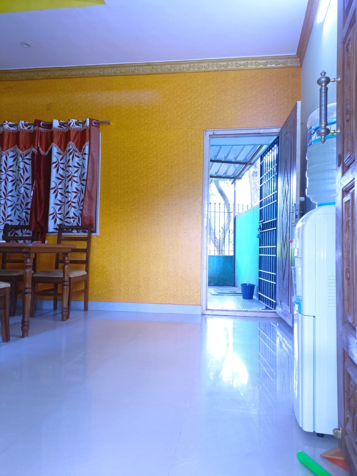The image captures a bright interior featuring a dining area with a wooden table and chairs. A vibrant yellow wall adds color to the space, while a door leads to an outdoor area. A water cooler and a small plant are also visible in the corner.