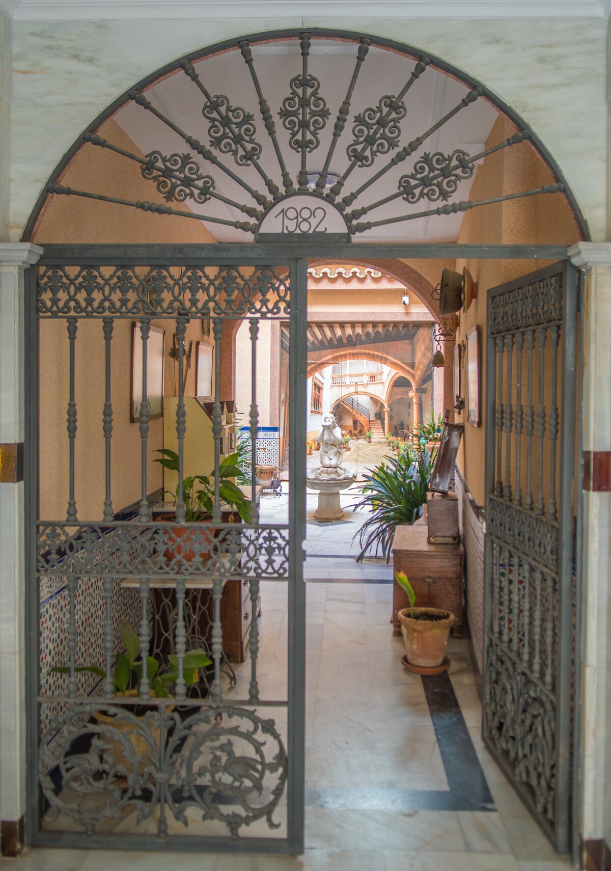 An ornate iron gate leads into a hallway adorned with decorative tiles and potted plants. Light filters through archways, illuminating a tranquil fountain at the far end, adding a serene touch to the inviting atmosphere.