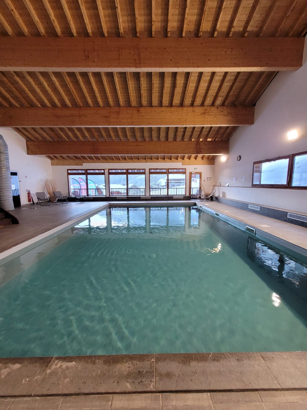 An indoor swimming pool is framed by large windows that allow natural light to fill the space. A wooden ceiling with exposed beams adds warmth to the area. The tranquil water surface reflects the ambient light, creating a calm atmosphere for relaxation.