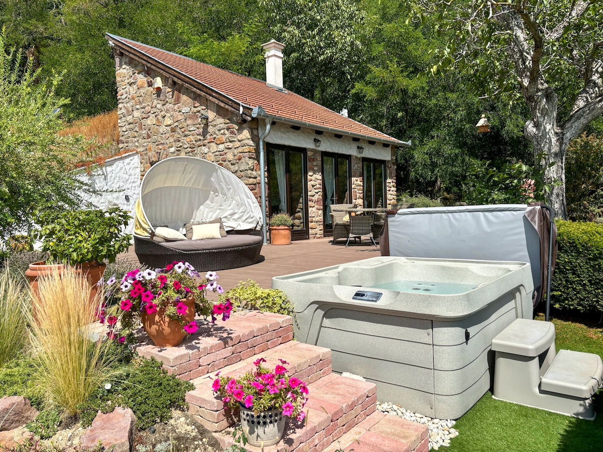 A charming stone cottage is set amidst lush greenery, featuring a spacious deck with a hot tub and seating area. Colorful flower pots add vibrancy to the scene, while natural stone steps lead up to the entrance, enhancing the cozy outdoor atmosphere.