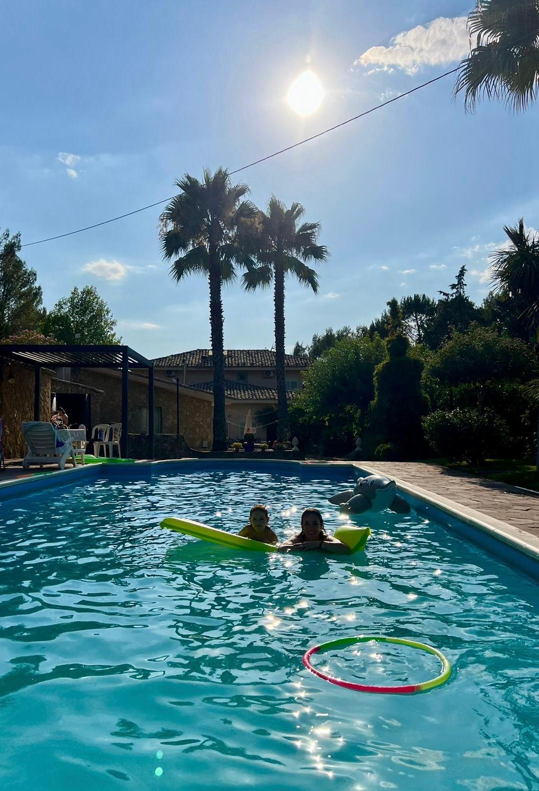 A sparkling pool reflects sunlight, surrounded by palm trees and lush greenery. A floating lounge and a colorful hula hoop are visible in the water. Guests can be seen enjoying the space, with shaded lounge areas present in the background.
