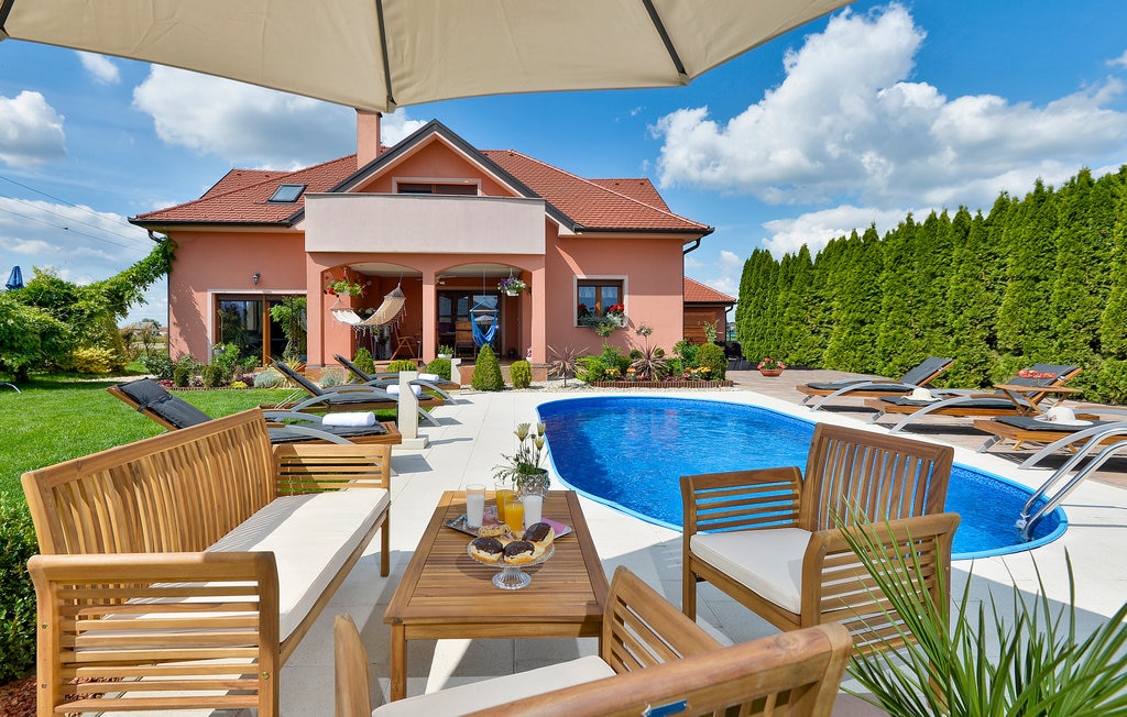 A spacious outdoor patio is featured, furnished with wooden seating and a table, set under a large umbrella. The inviting pool area is surrounded by sun loungers and lush greenery, while the home's exterior is shown in a charming design with large windows.